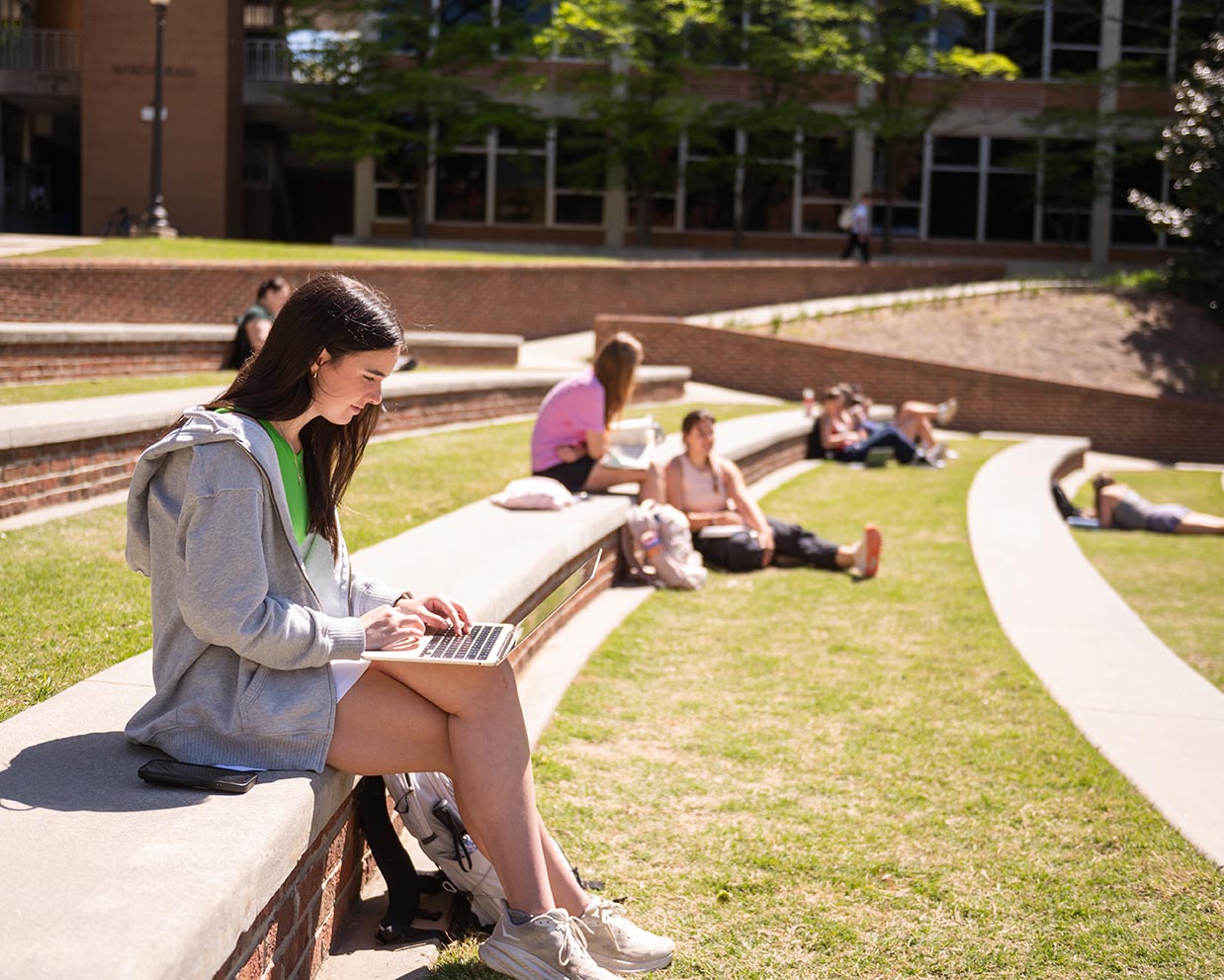 Student sits on a curved outdoor bench using a laptop in a sunny campus amphitheater, with other students relaxing on the grass behind her