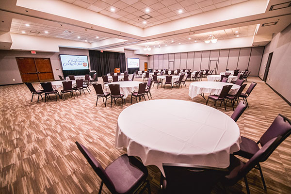 A spacious banquet hall set up for an event, featuring circular tables with white covers and a stage in the background.