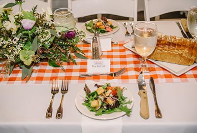 A beautifully set dining table featuring a fresh salad, water and wine glasses, a loaf of bread, and a floral centerpiece on a checked tablecloth.