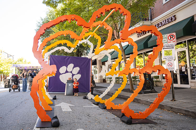 A young boy poses with a replica of Howard's Rock behind a series of four large metal figures that depict the outline of the Clemson Tiger Paw.