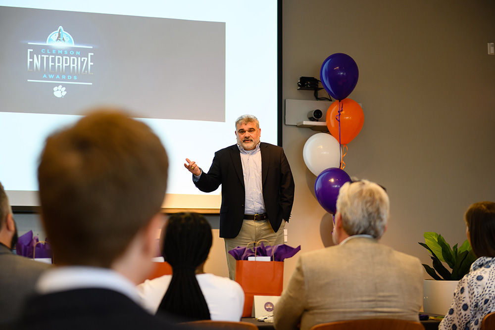 A speaker presenting at the Clemson EnterPrize Awards event, with a logo displayed on screen and an audience in view.