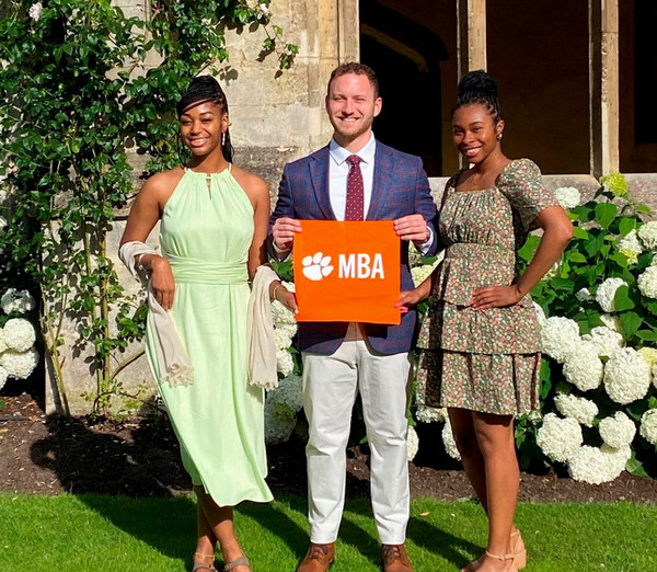 Two women and a man pose for a photo holding an orange "MBA" sign, surrounded by greenery and flowers.