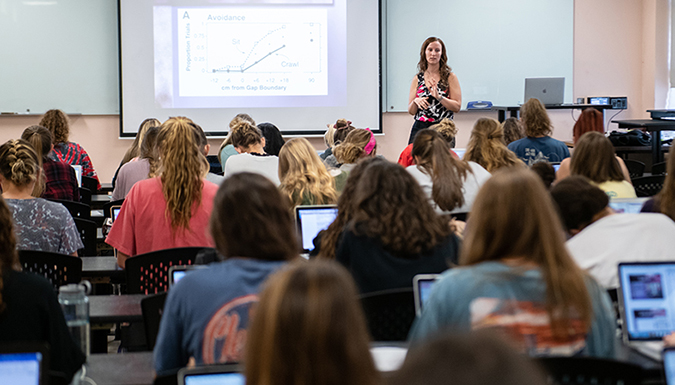 Several students facing the projection of a graph on the board, take notes on laptops during a lecture in a psychology course.