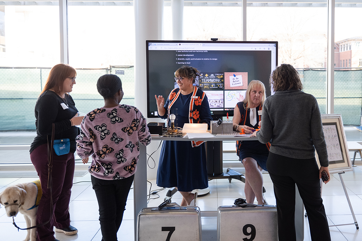 People chatting at an information booth with a presenter speaking in front of a large screen display.