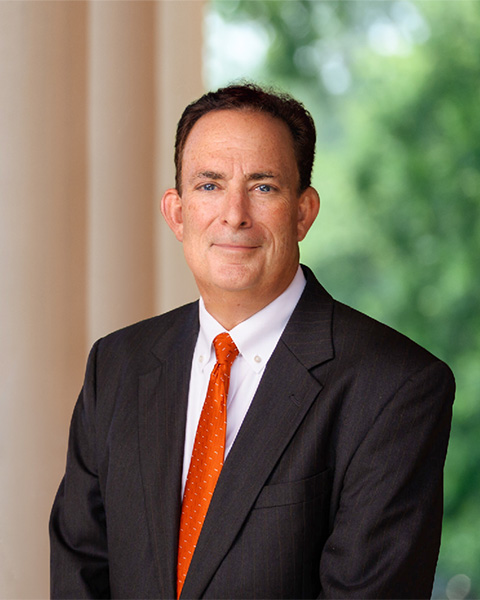 The President in a dark suit with a white shirt and bright orange tie, standing outdoors with blurred greenery in the background.