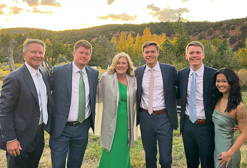 The Guskiewicz family dressed in formal attire pose outdoors at sunset, with autumn trees and rolling hills in the background.