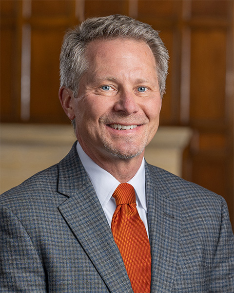 A smiling President-Elect Guskiewicz in a gray suit with an orange tie in an indoor office setting.