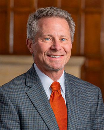 A smiling President-Elect Guskiewicz in a gray suit with an orange tie in an indoor office setting.