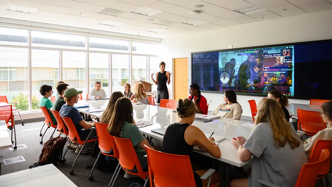 A classroom setting with students seated around a table, engaged in a discussion with a presenter in front of a large screen displaying multimedia content.