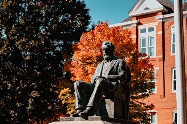 Bronze statue of Thomas Green Clemson seated against a backdrop of vibrant autumn foliage and Tillman Hall, a historic brick building.