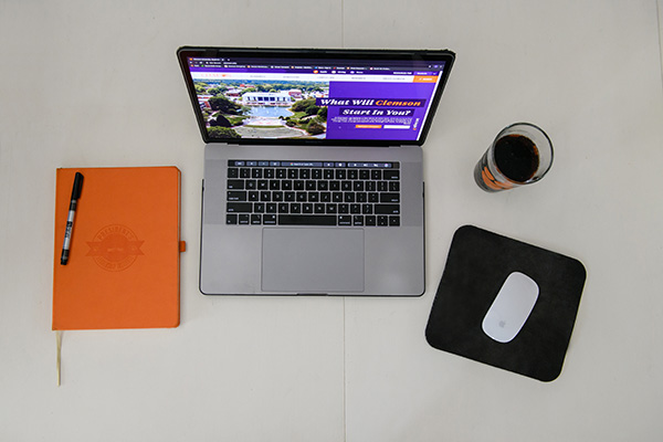 Laptop with Clemson's Website displayed on the screen sitting with a mouse, an orange notebook and a Clemson cup on a white desk. 