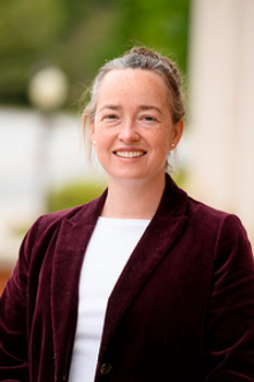 Headshot of a smiling woman with light brown hair in a bun, wearing a burgundy blazer and a white shirt, set against an outdoor background.