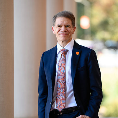 Provost Smith stands outdoors, smiling in front of columns. His tie features a colorful paisley design, and he wears an orange tiger paw on his lapel.