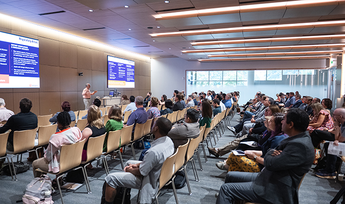 New faculty members sitting in conference style room listening to presenter speak with two large screens to either side of them.