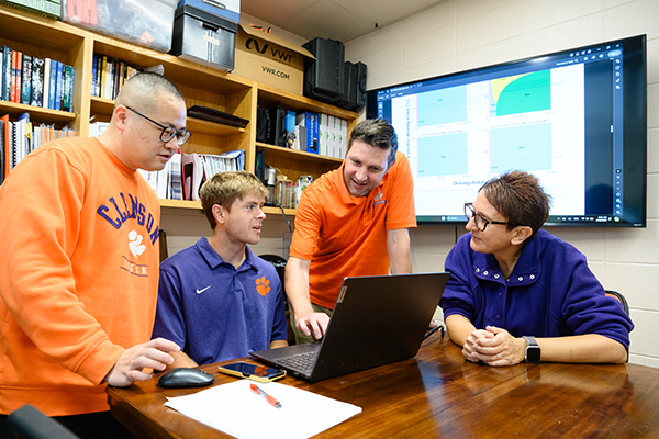 Professor with three students collaborating around a laptop in a study room, reviewing data displayed on a large screen.