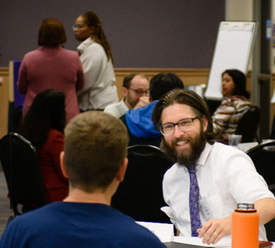 A man with glasses and a tie smiles while engaging with a participant in a workshop setting, with other attendees conversing in the background.