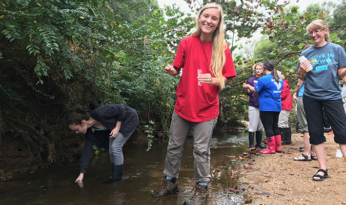 A volunteer smiling, standing in a stream, holding pH test tubes as another volunteer kneels to collect samples. Trainer Carrie and other volunteers stand on the beach.
