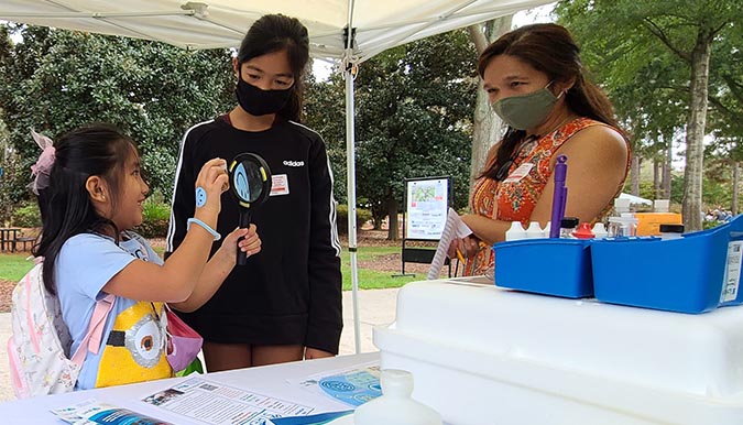 A child holds a sample tube and magnifying glass for two other people to see, while standing next to a table with SC Adopt-a-Stream brochures and supplies on display.