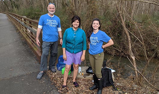 Trainer Madeleine standing with two people next to the Reedy River, with water sampling supplies on the ground next to them.