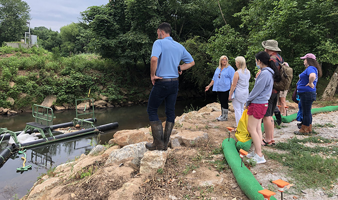 A group of people standing next to the Reedy River looking at a Bandalong Litter Trap while a representative from ReWa explains how it works.