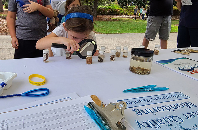 A child using a magnifying glass to look at macroinvertebrates in sample bottles on a SC Adopt-a-Stream display table.
