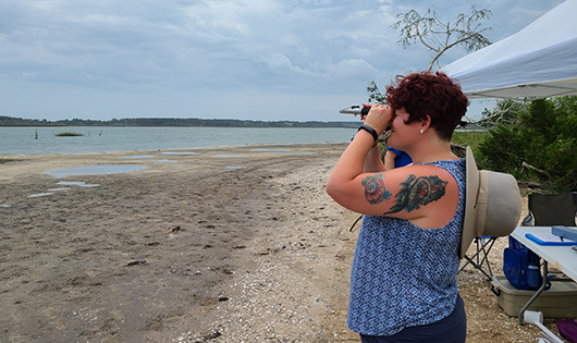 Trainer Cara standing on a beach facing the water as she looks into a refractometer.
