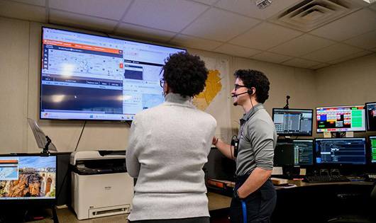 A male wearing a headset and a female in a room with computers and a big digital screen looking at data