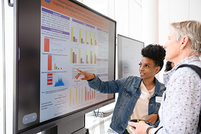 Female graduate student points at large digital board displaying graphs. 