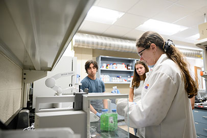 Students conducting experiments in a laboratory setting, with one student wearing a lab coat and handling equipment while others observe.