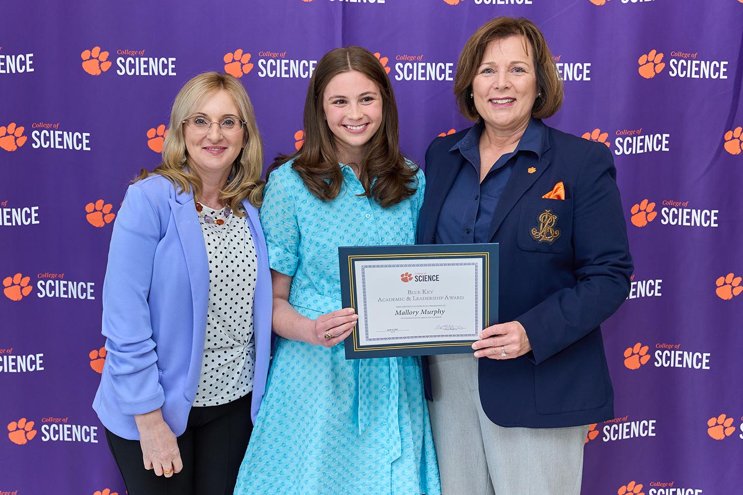 Three women stand in front of a purple banner with the College of Science logo stepped and repeated across it. The middle woman is holding an award plaque.