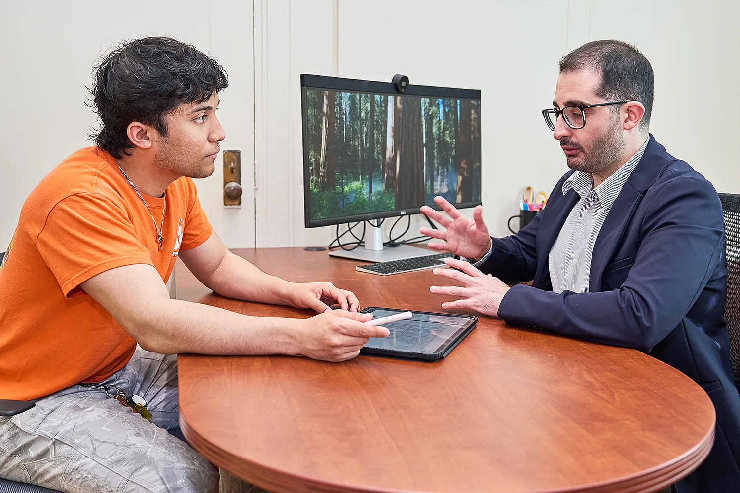 Two people talk at a desk in an office with a tablet and computer display.