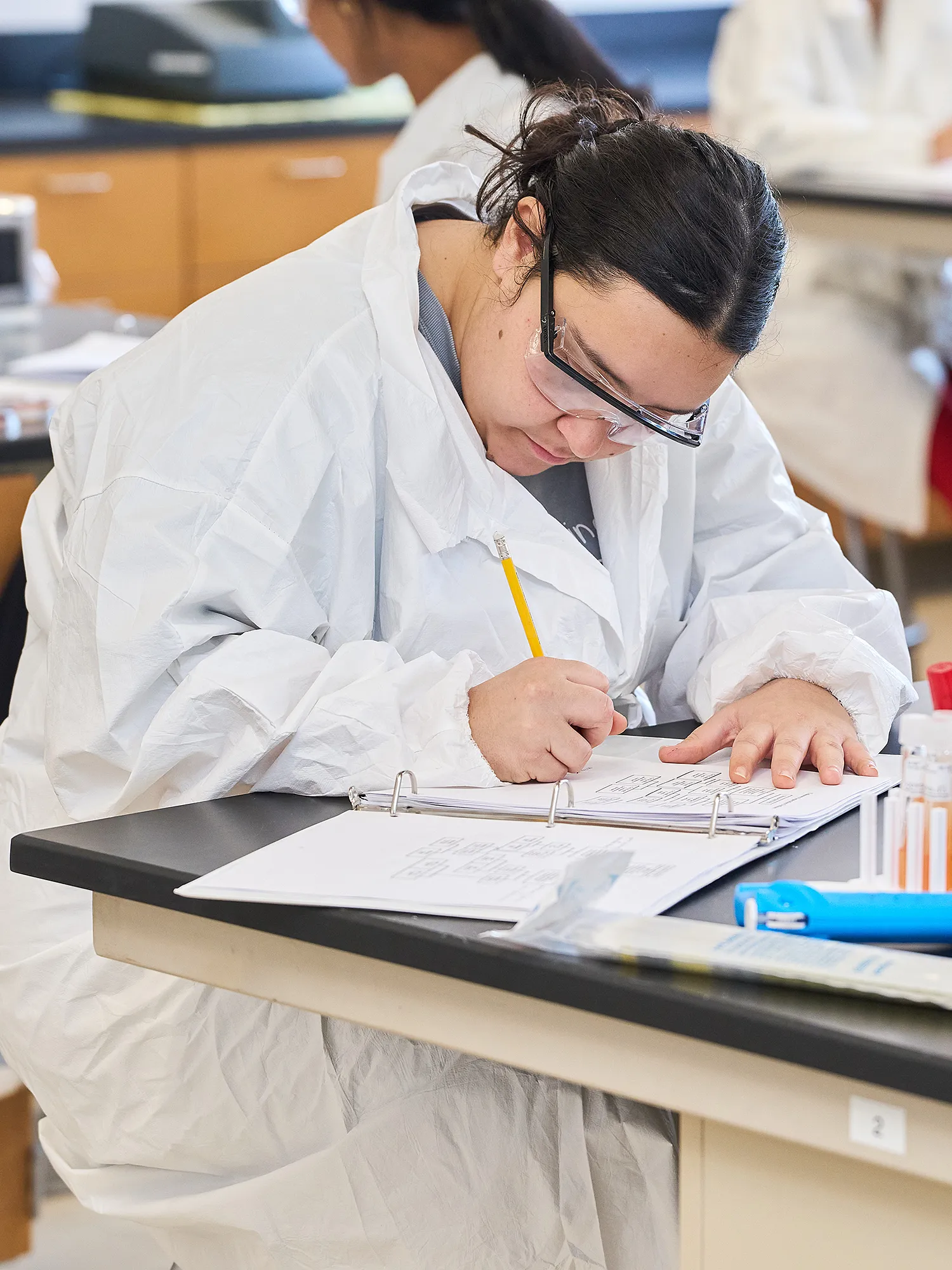 Student wearing lab coat and goggles taking notes in a lab.