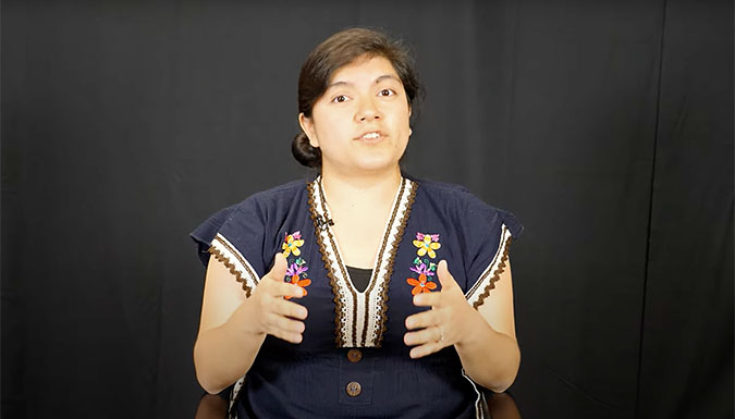 Female student speaking to camera during interview, with black drape behind her.