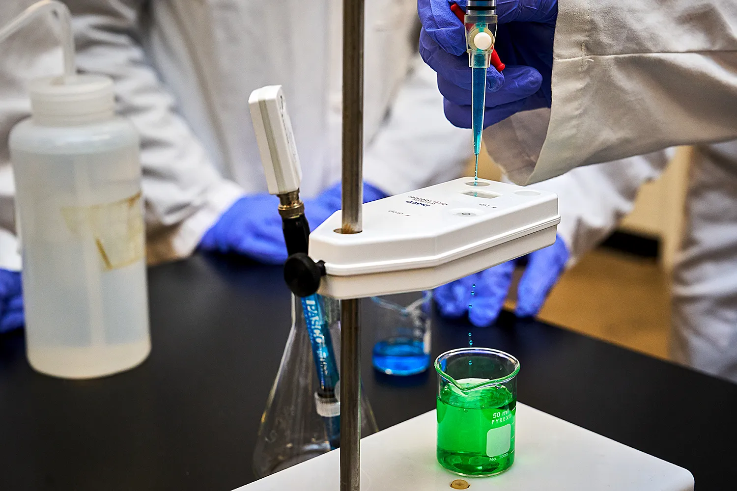 Gloved hands adjusting a dropper, adding green liquid to a beaker below.