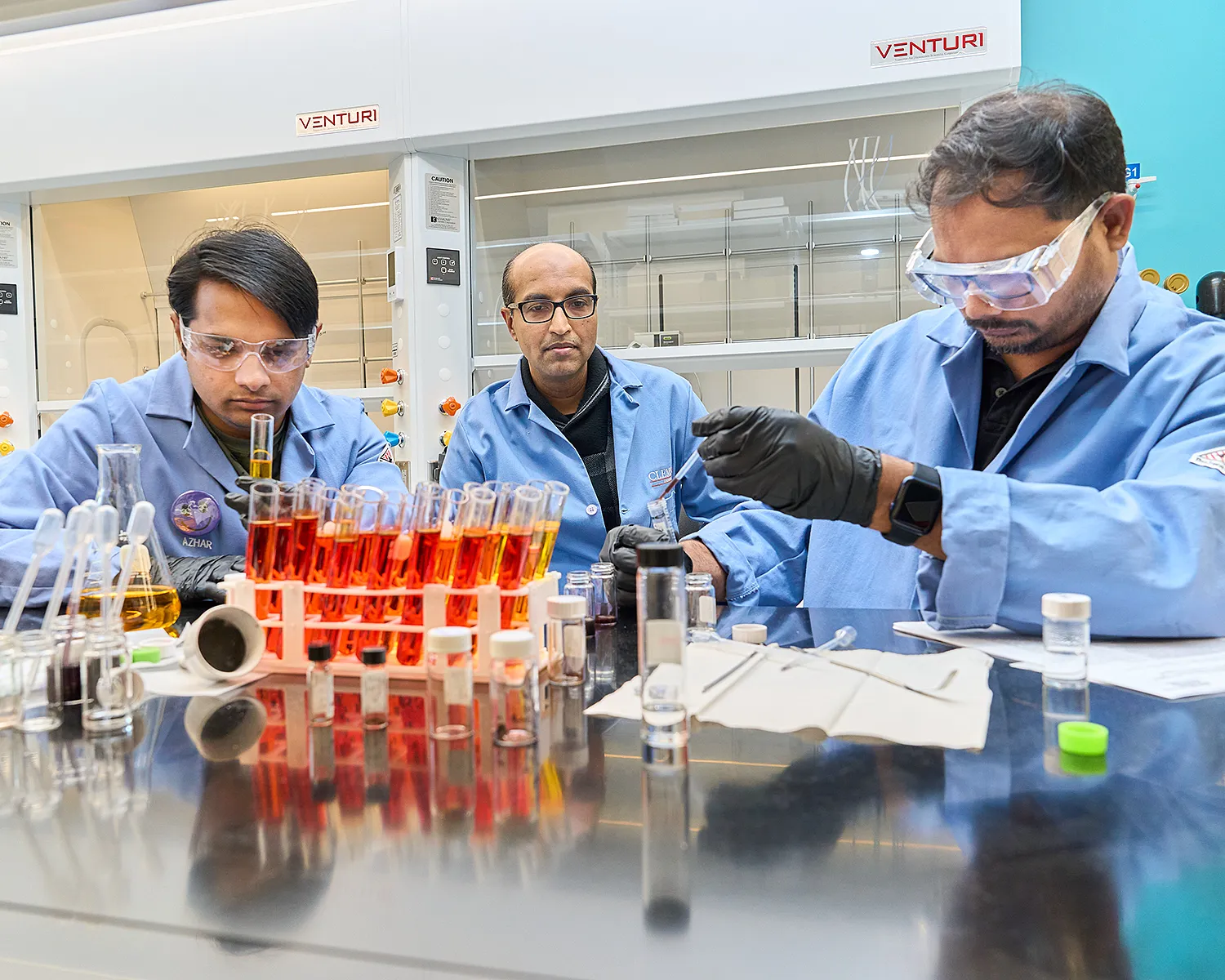 Three men working in a science lab, wearing blue lab coats, with orange test tubes in front of them.
