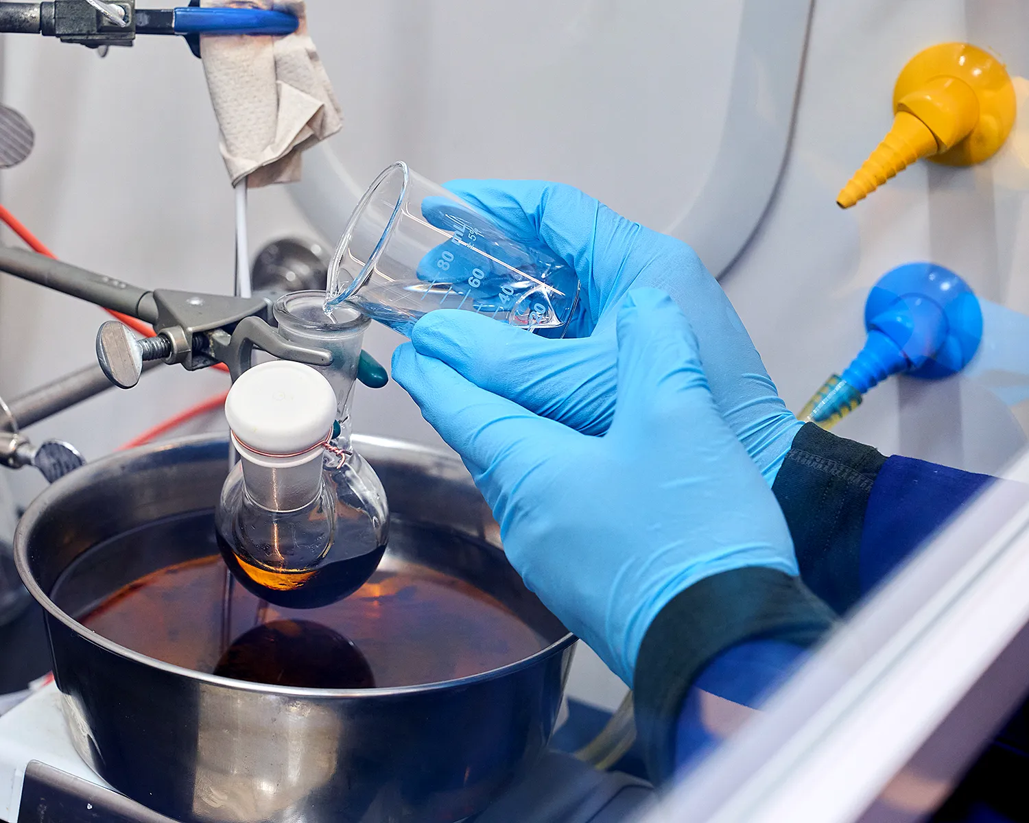 A blue gloved hand holding a small beaker and pouring its contents into another beaker suspended over a heat bath.