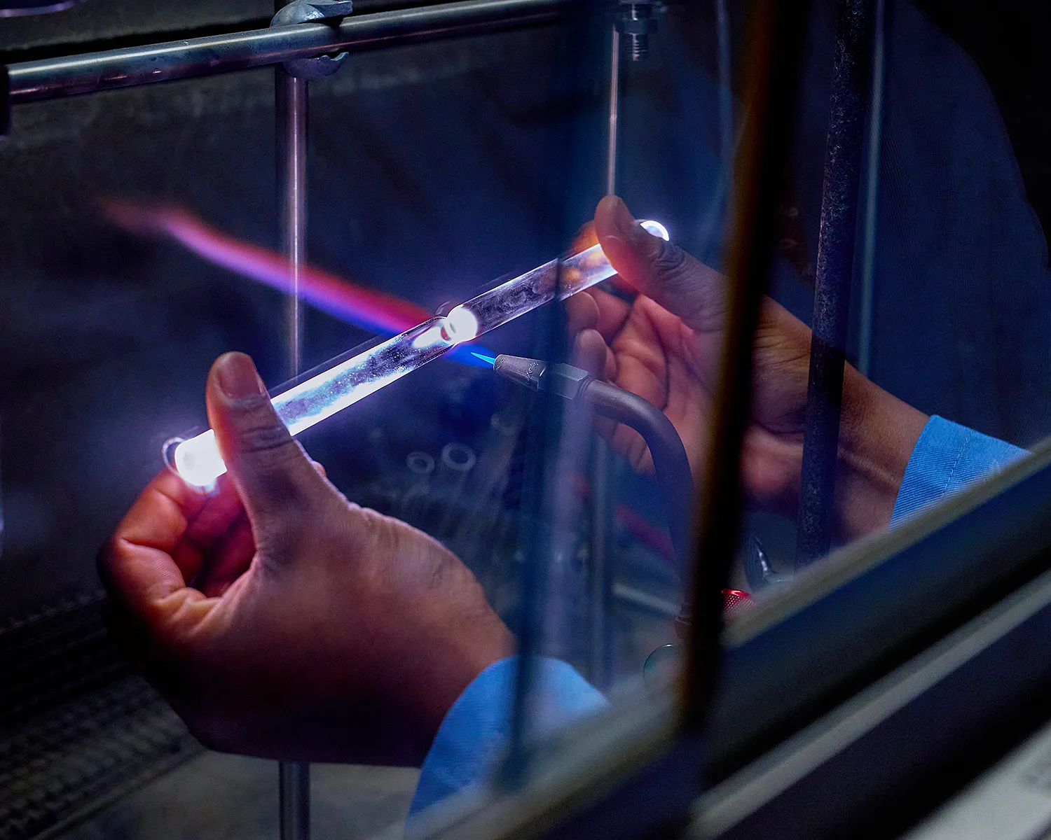Two hands holding a glass tube being cut by a torch, with blue and orange flames running through the glass.