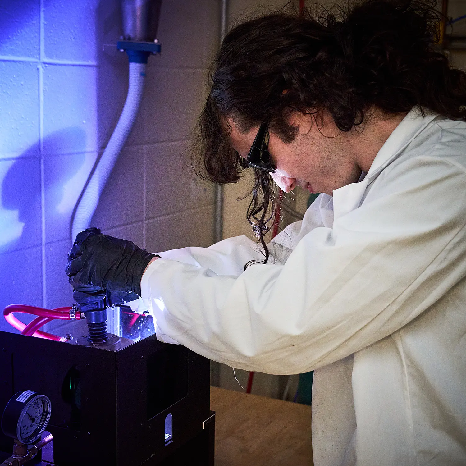 Man, with goggles, gloves and lab coat working in lab with a box containing bright bluish light.