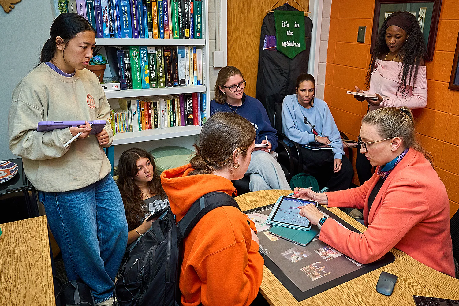 A person at a desk uses a tablet to explain something to a person, while five others sit and stand around the desk watching.