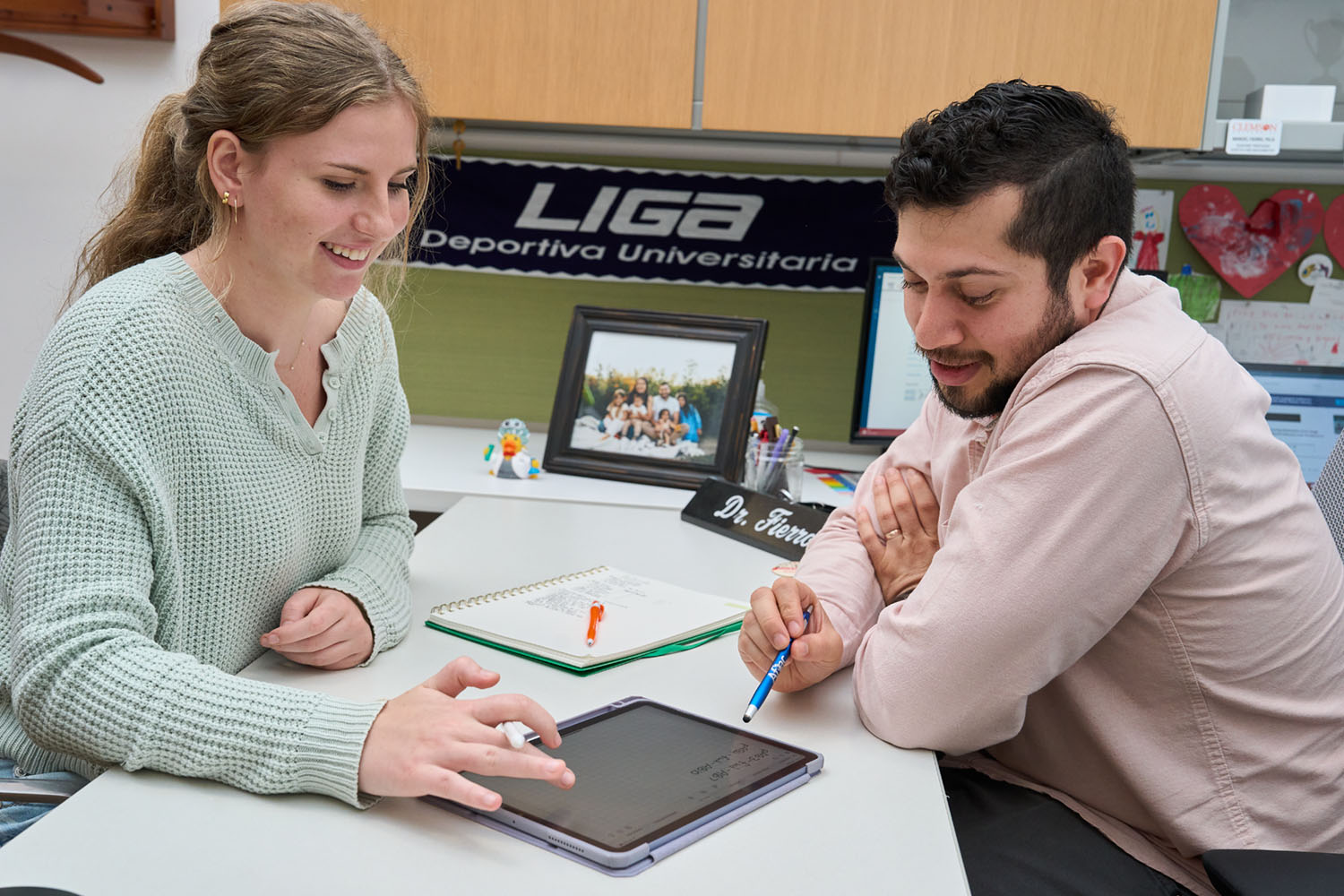 Student speaking with professor in office, working on tablet.