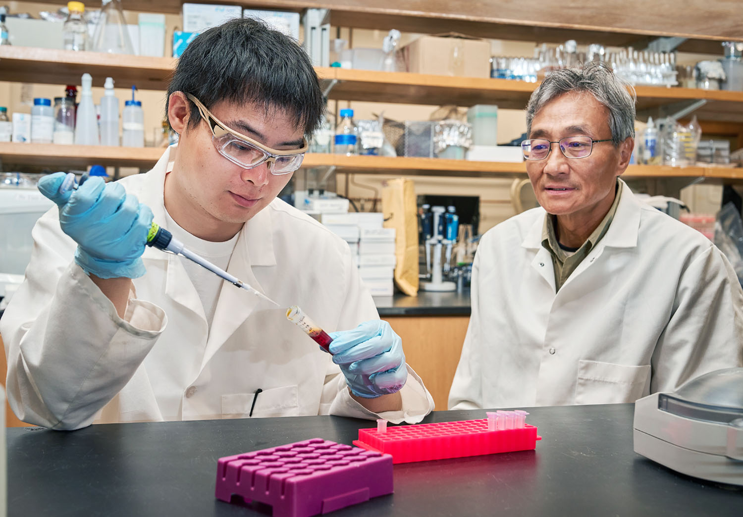 Grad student working in lab pipetting a red liquid while a professor looks on.