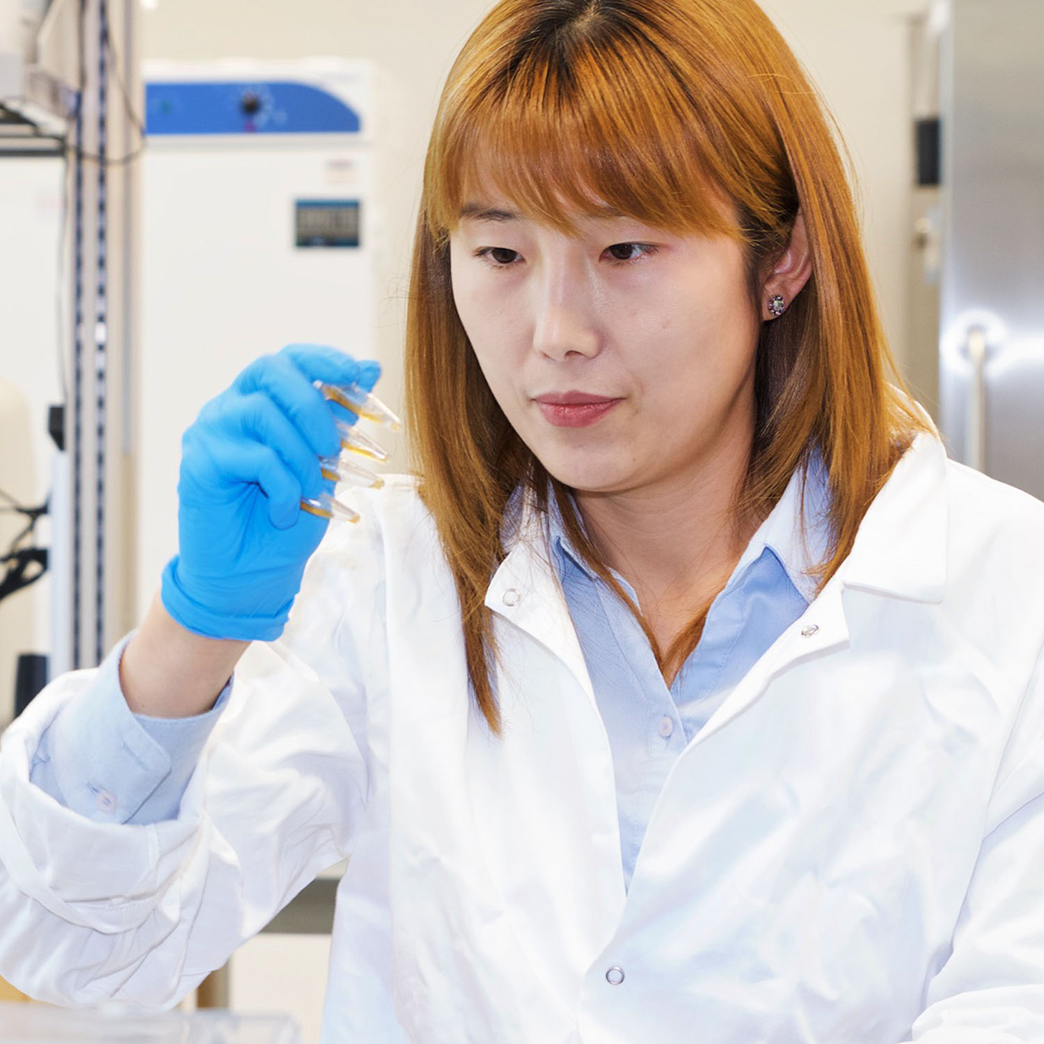 Woman in white lab coat in lab, with blue gloves, holding three small vials.