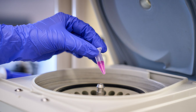 Blue-gloved hand holding sample vial with purple liquid, being placed into centrifuge.