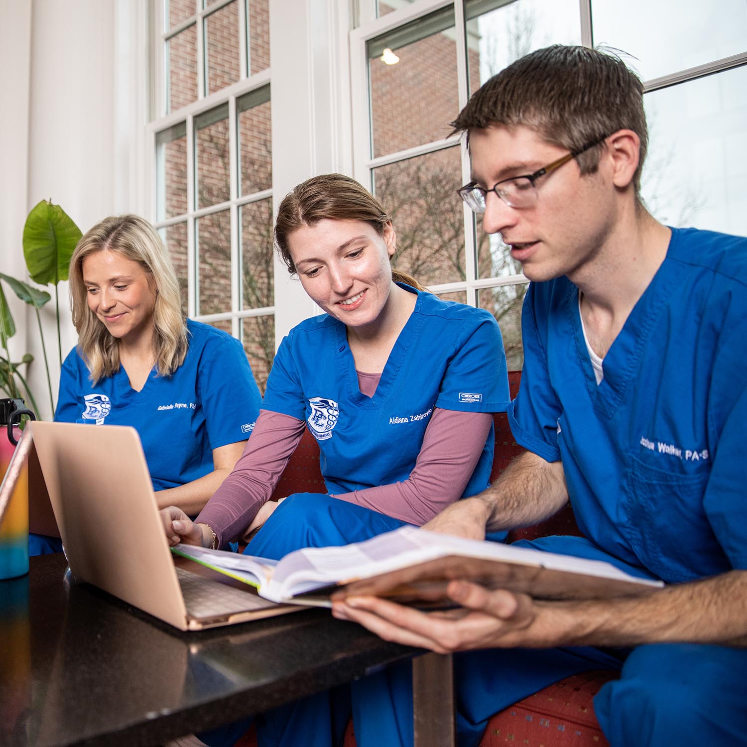 Two female medical students and male student, all wearing blue scrubs, working on a laptop PC.