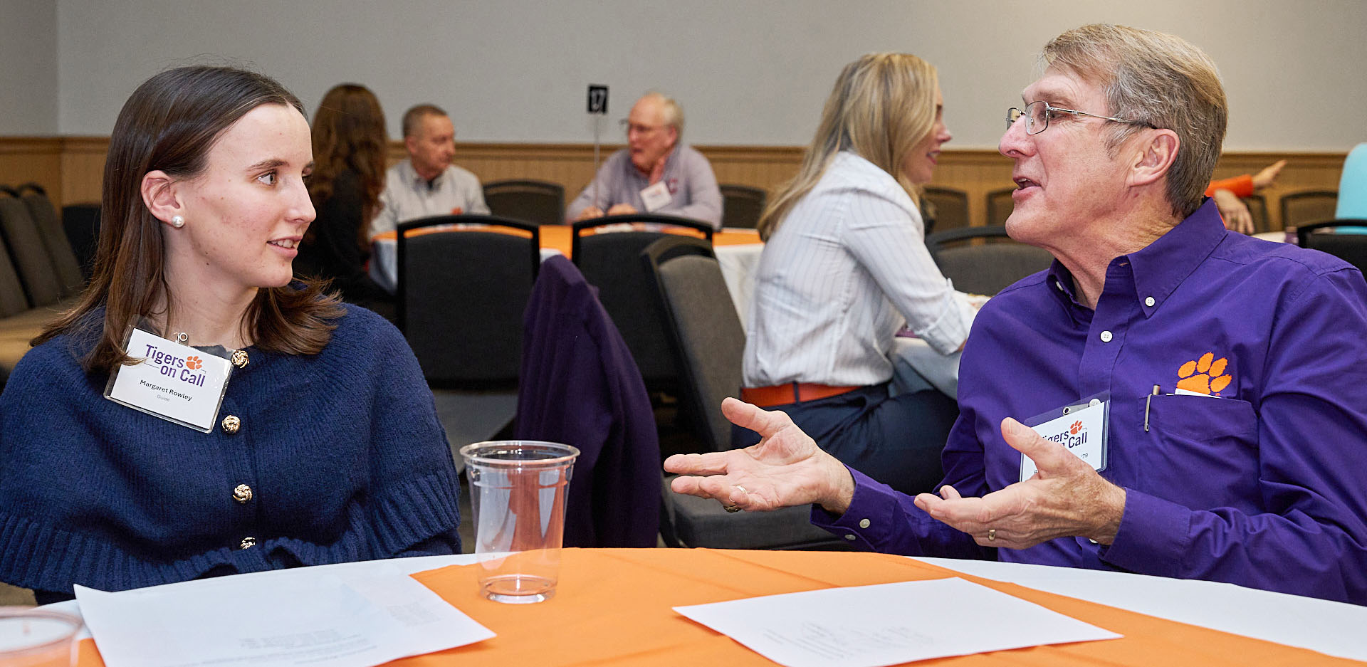 A woman and man sitting having a roundtable discussion, with other people at tables in background.