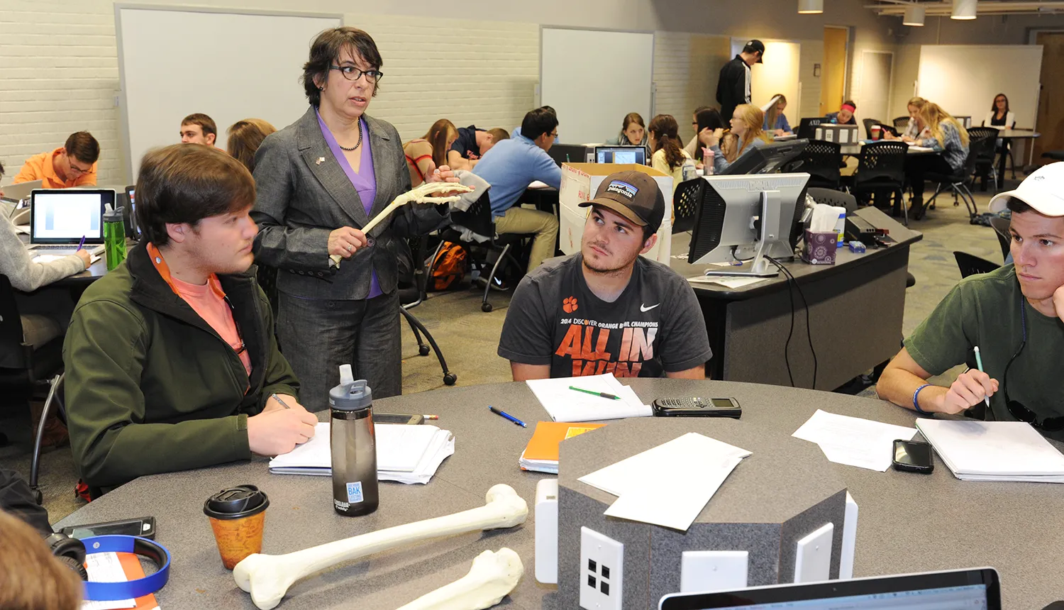 A female instructor speaks to three students sitting at a round workstation.