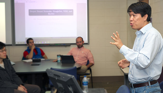 An instructor speaking to a class of students in a classroom, with blurred images projected on screens.