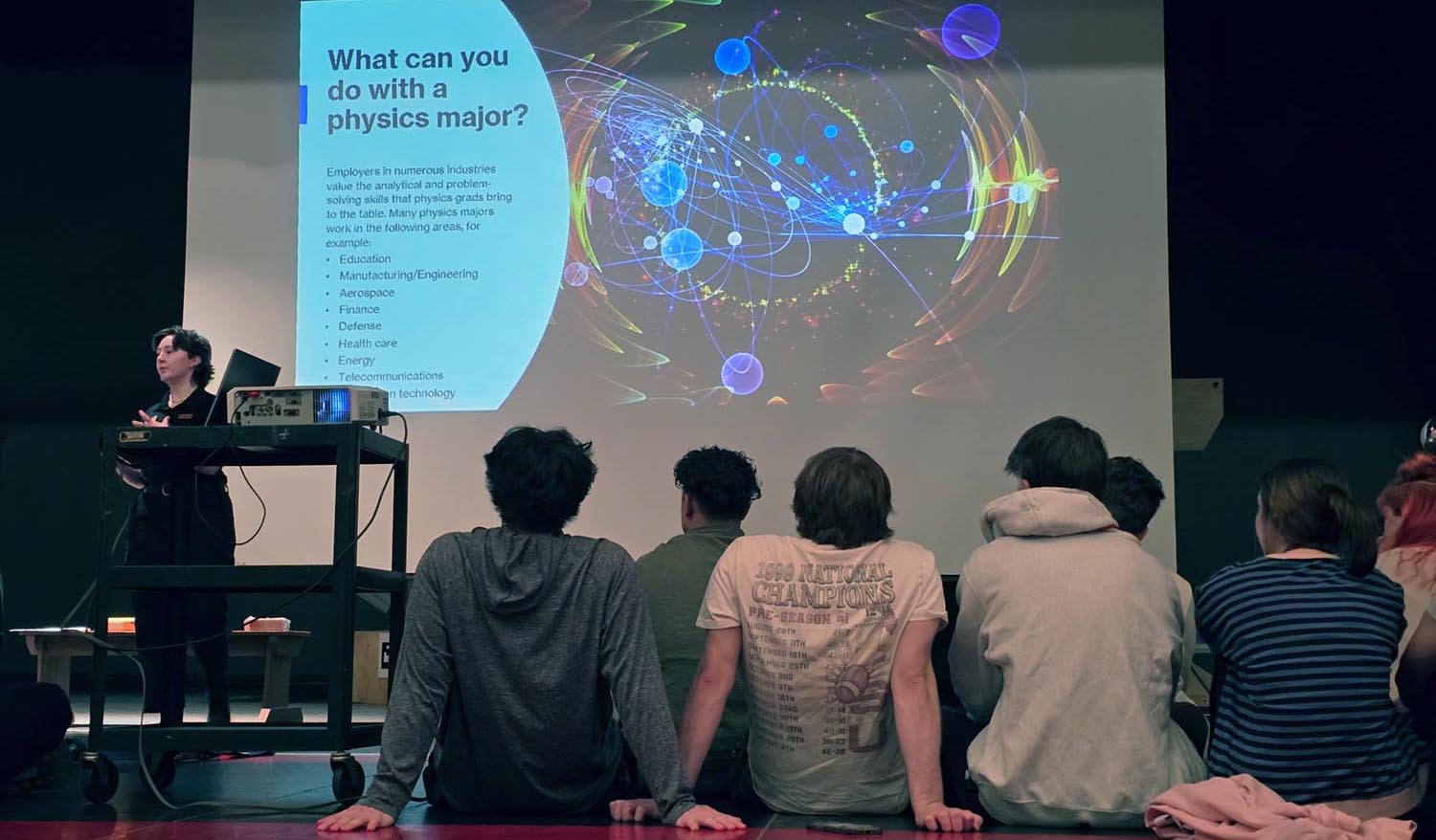 Person at podium on left speaking to group of children, with their backs toward the camera. A presentation is displayed on an overhead screen.