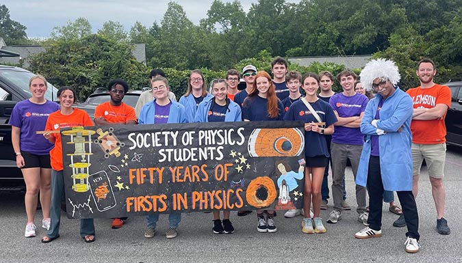 Group of people in a parade, some in costumes, holding a sign, Society of Physics Students - 50 years of firsts in physics.