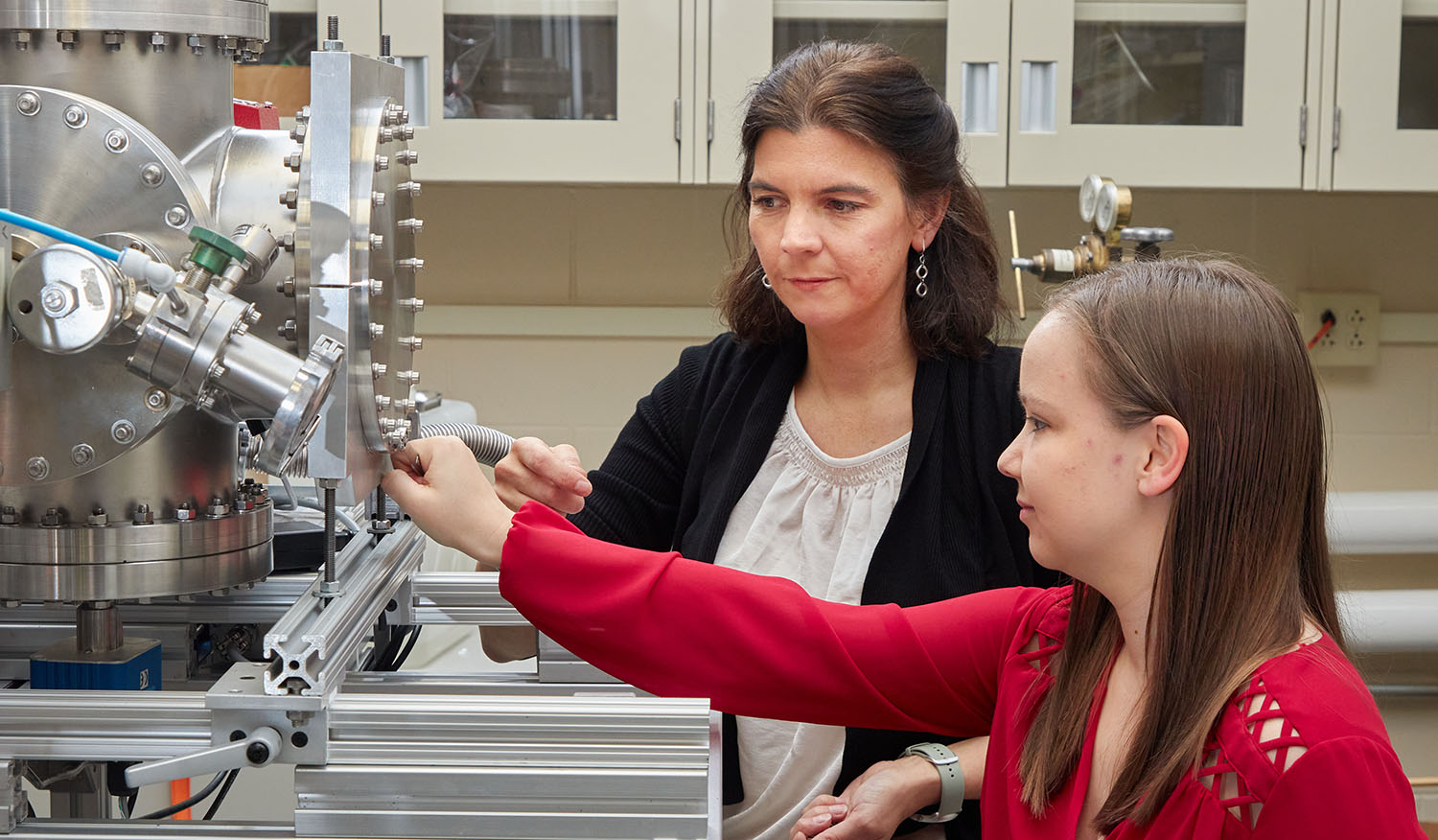 Two women work together to adjust a large scientific instrument in a laboratory, with one pointing at a control while the other observes.